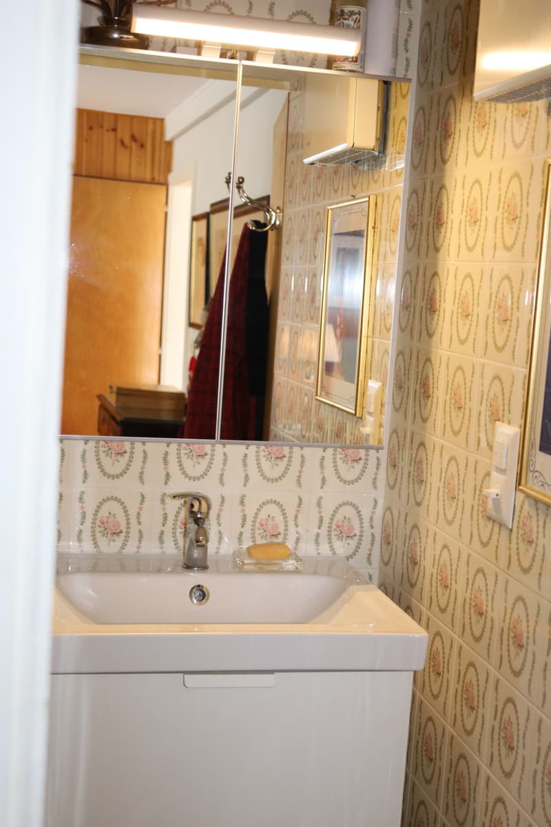 Small bathroom with white ceramic sink, mirror, tiled wall with flower pattern, white ventilation grill, white switch on wall