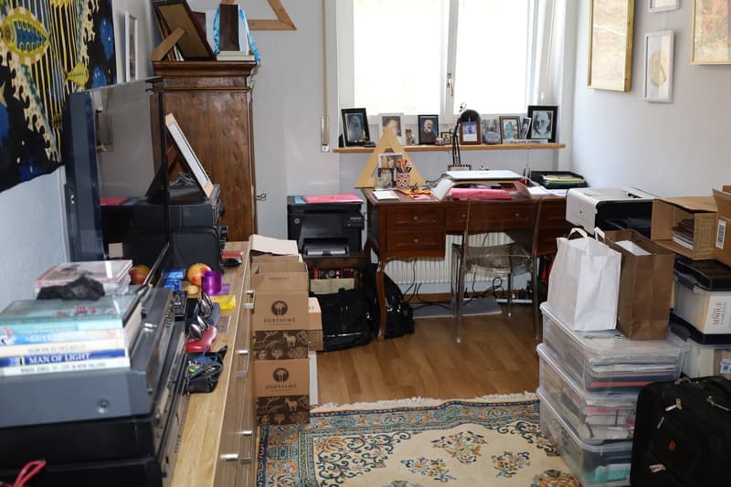 A room with a desk, chair, boxes, printer, radiator, and storage boxes.