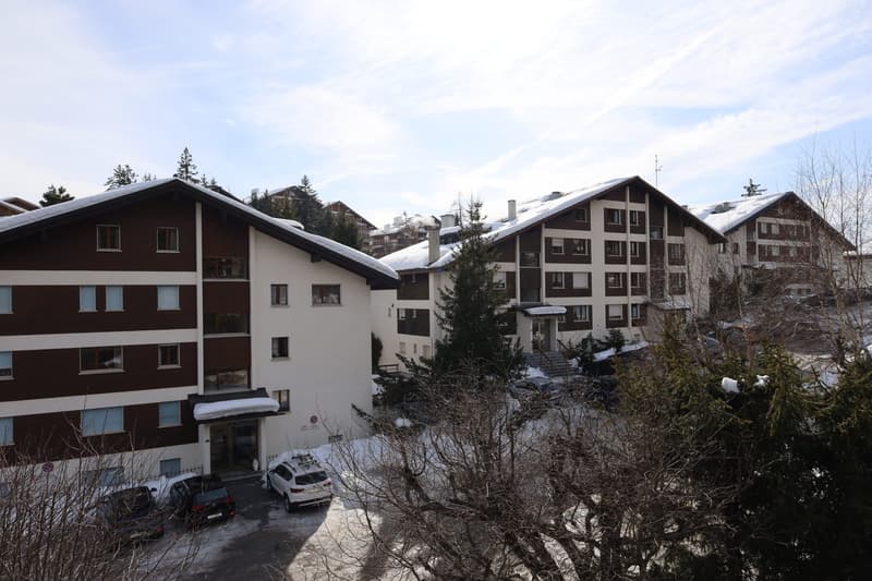 A multistory apartment building with a parking lot, covered in snow, located in a ski resort area, surrounded by pine trees.