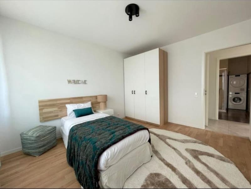 Spacious bedroom with a wooden headboard, white bedding, and a patterned blanket. There is a wardrobe and a washer/dryer visible in the room.