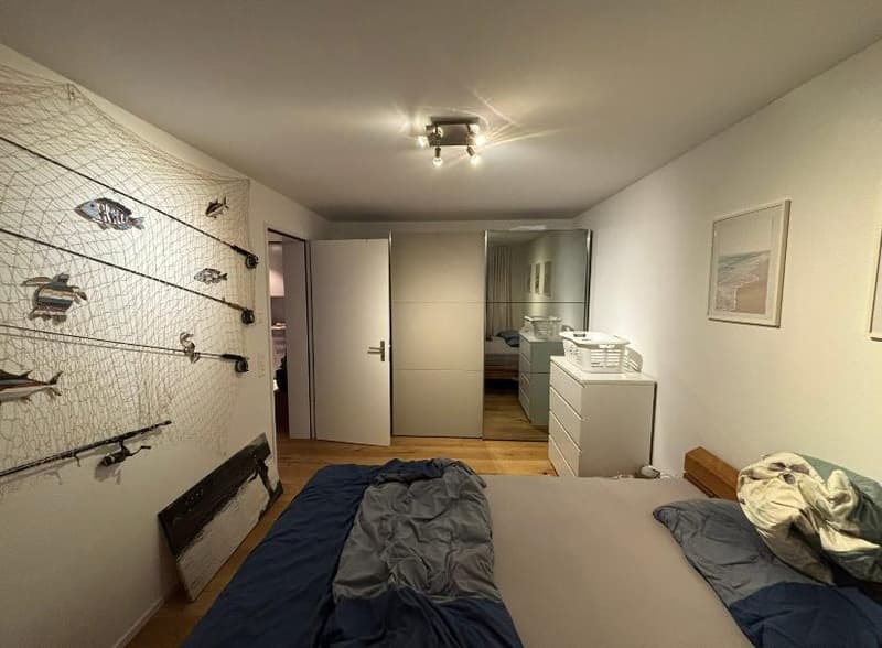 Minimalistic bedroom with a bed, open door, white walls, decorative net with fish pictures, ceiling lights, wooden floor, chest of drawers, mirror