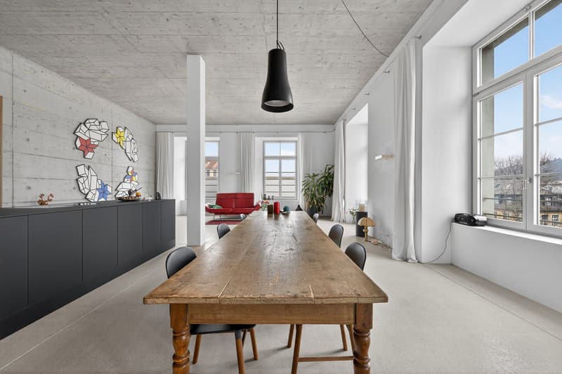 large room, long wooden table, black chairs, black cabinets, white walls, large windows, red couch in the other room