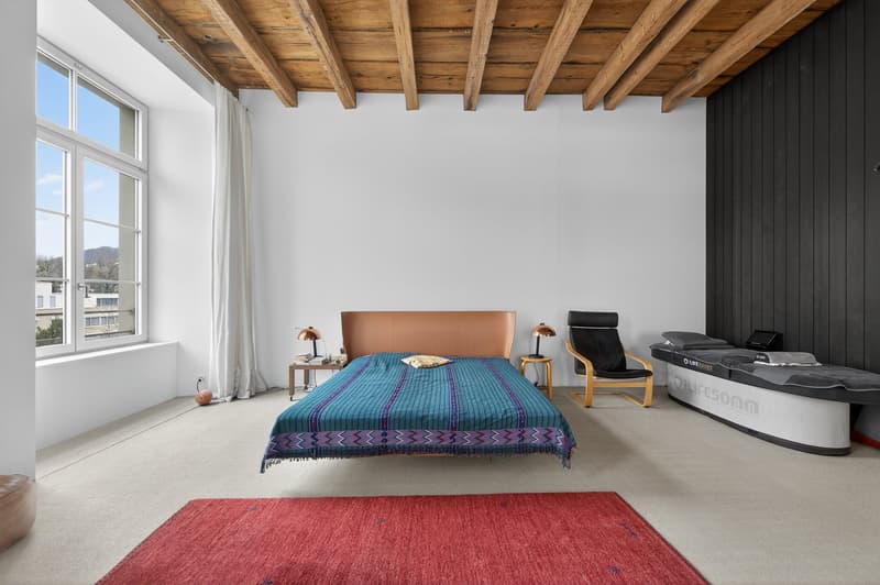 Spacious bedroom with wooden ceiling, a bed, two bedside tables, a nightstand with a lamp, a chair, and a massage table. There is a red rug on the floor, and a window with a view of the outside.