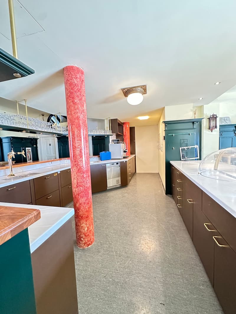 Modern kitchen, large kitchen island, central pillar, stainless steel appliances, wine glasses on a shelf, green cabinets, white countertops, ceiling light