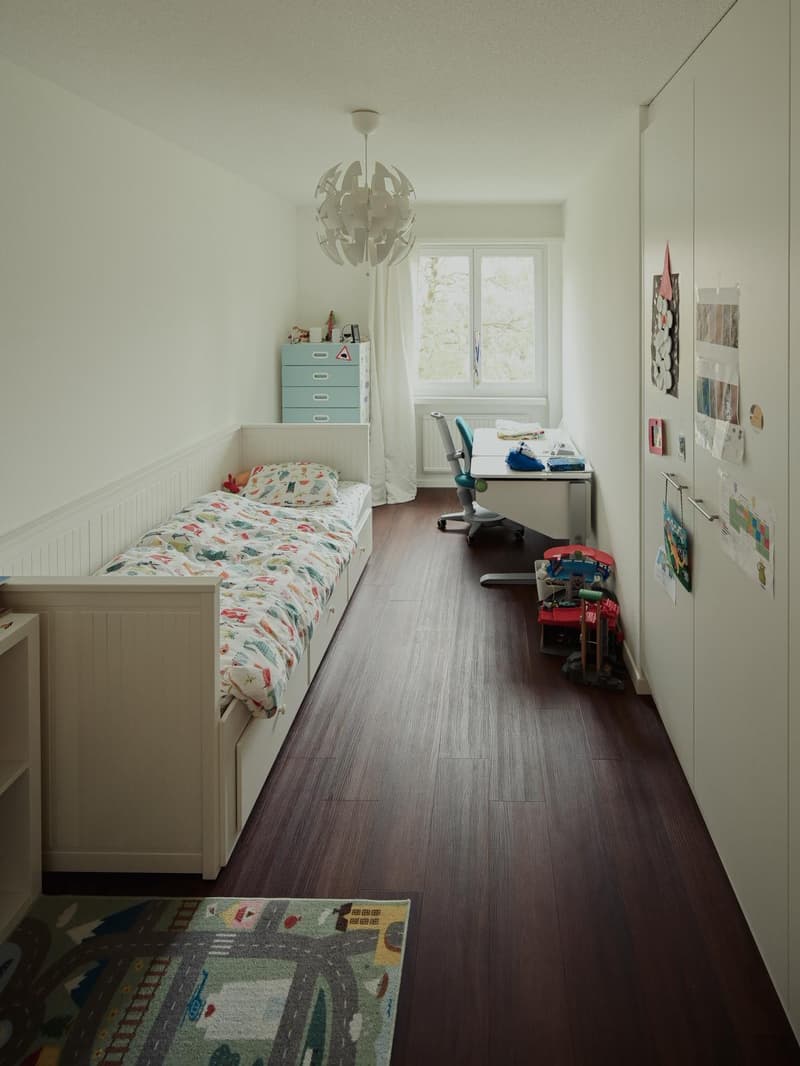 single bed, blue drawers, white furniture, wooden floor, white walls, window with curtains, small rug, small desk with chair, toy blocks, lamp