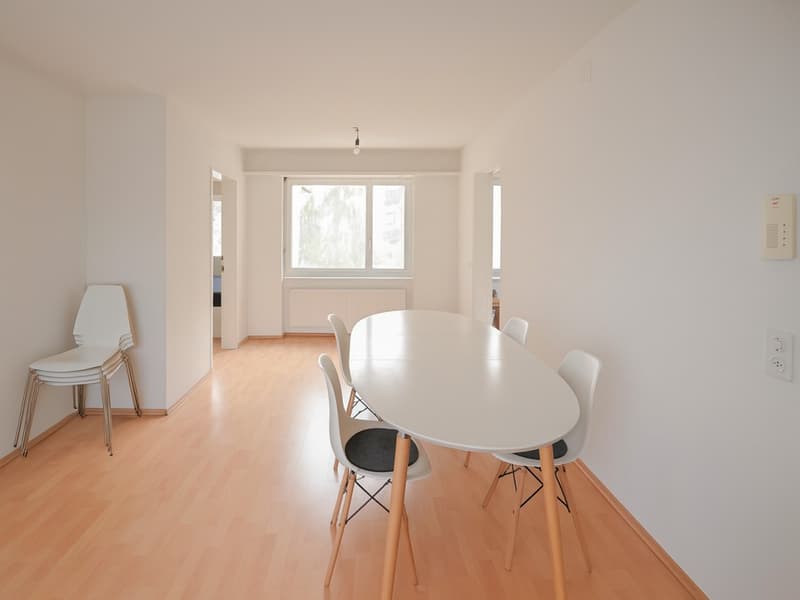 an empty room with a long table and chairs, wooden floor, white walls, small window
