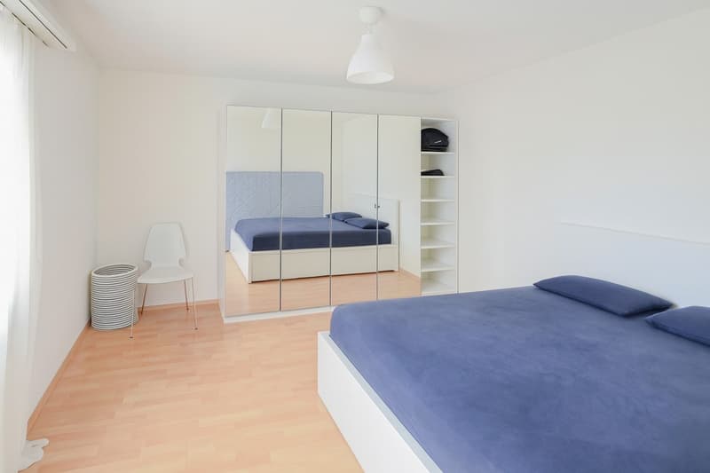 bedroom with white walls and flooring, large mirror, large bed, blue bedding, shelf, chair, round light fixture