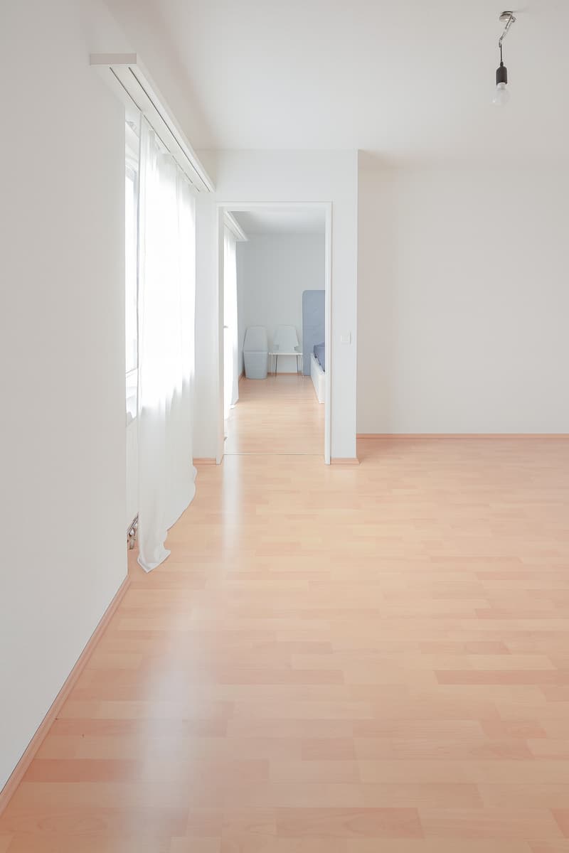 Open, unfurnished room with wooden flooring and white walls. Interior doors lead to another room, which has a bed, chairs, and storage bin.