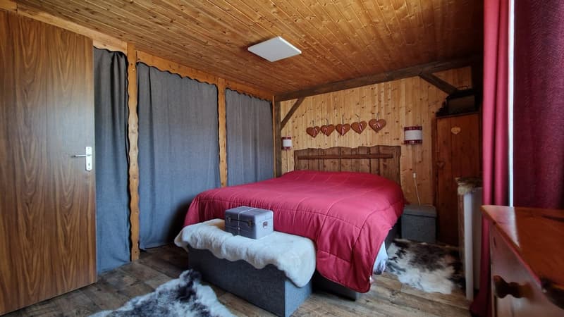 Cozy wooden cabin bedroom with a large red comforter on the bed, wooden walls and ceiling, and decorative heart-shaped wall hangings. There is a small chest or trunk at the foot of the bed, and a fur rug on the floor.