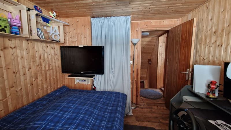Cozy wooden bedroom with a TV, shelves, and a curtain separating the room. The room has a blue plaid bedspread and a wooden floor.