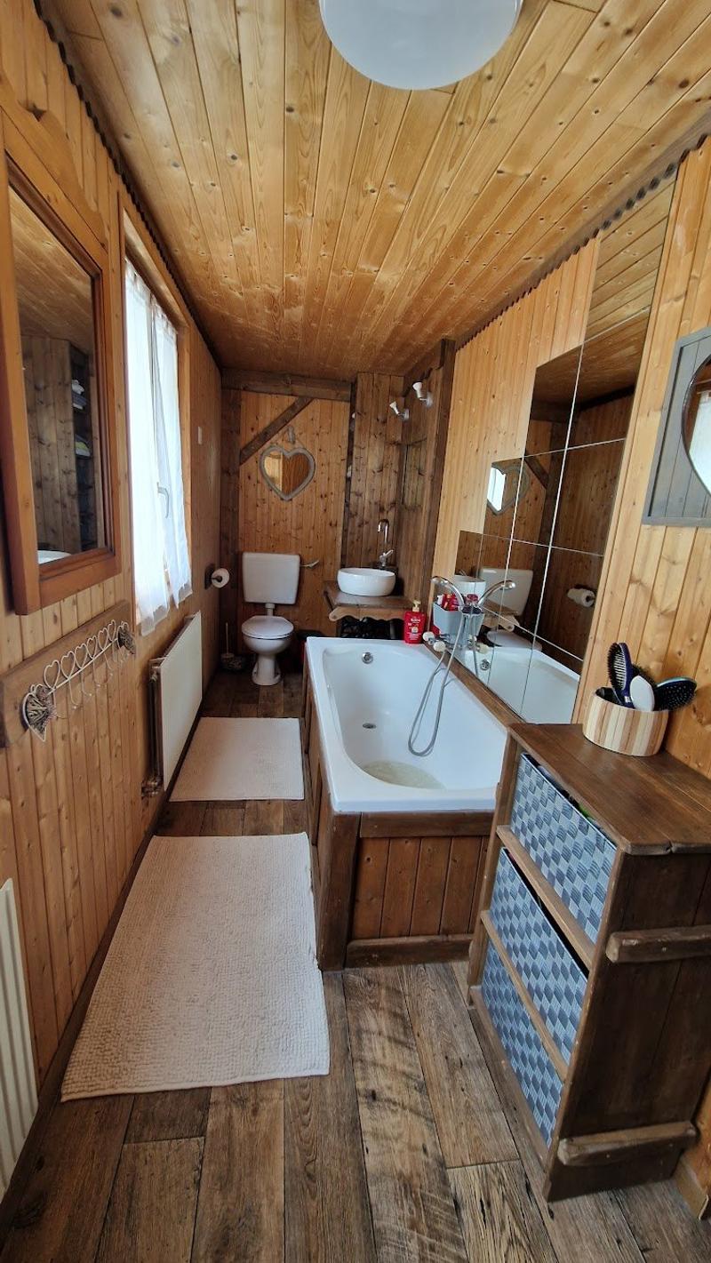 The image shows a rustic bathroom with wooden walls and ceiling, a bathtub, a toilet, a sink, and various bathroom accessories such as a mirror and storage units.