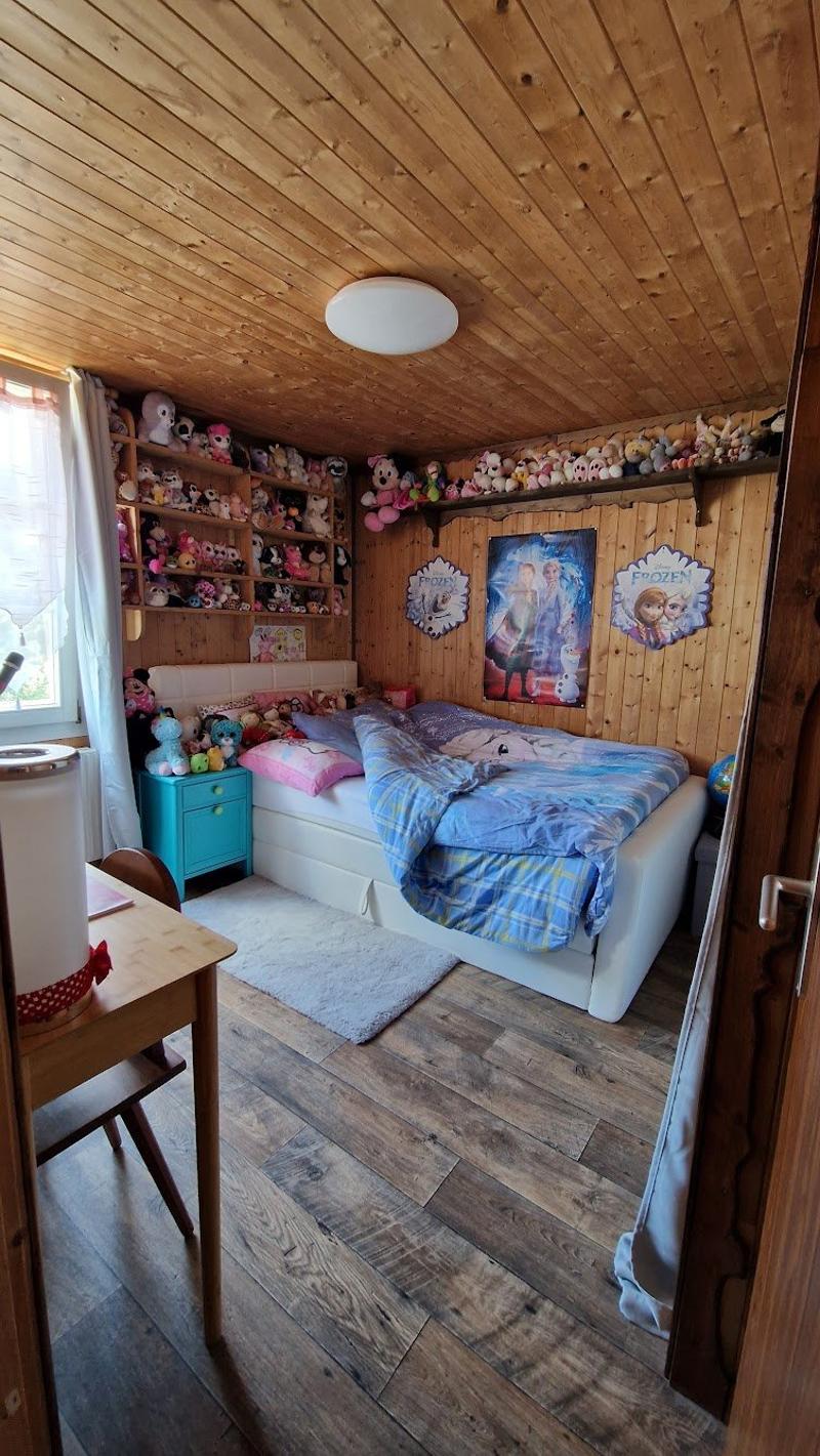 Cozy bedroom with wooden walls and ceiling, plush bedding, shelves filled with stuffed toys, and wall decor featuring Disney characters.