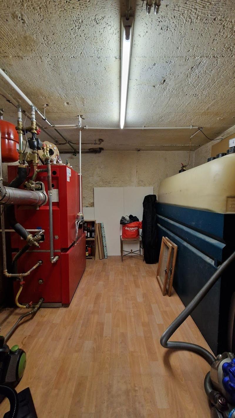 Wooden floor, white walls, pipes and valves on the wall, red storage box, shelves with wine bottles, a red bag and a black bag on the floor, and a white box.