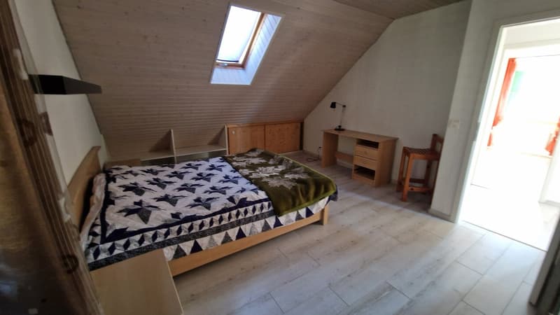 Bedroom with a bed, bedside shelves, wooden desk, and a small wooden chair. The wall has a sloped ceiling with a skylight window.