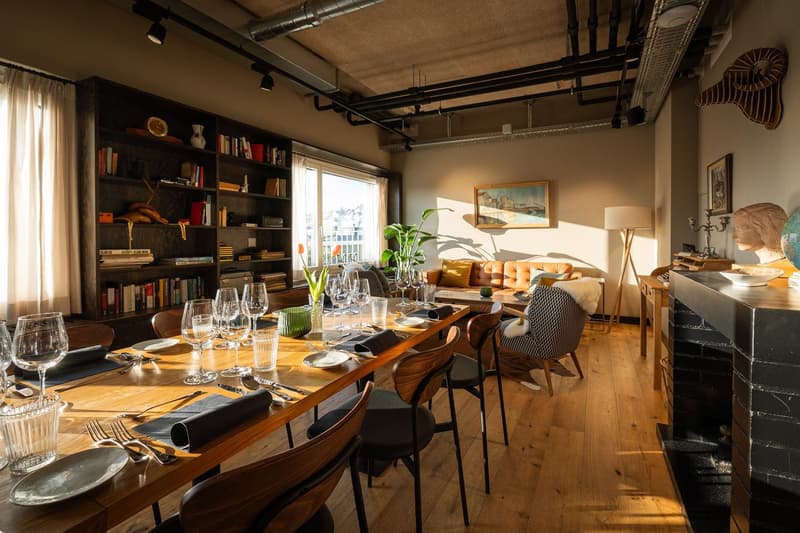 Spacious dining area with a large wooden table, black chairs, and a cozy fireplace. The room features exposed ceiling beams, bookshelves, and artwork on the walls. The space has a warm, industrial-chic ambiance.