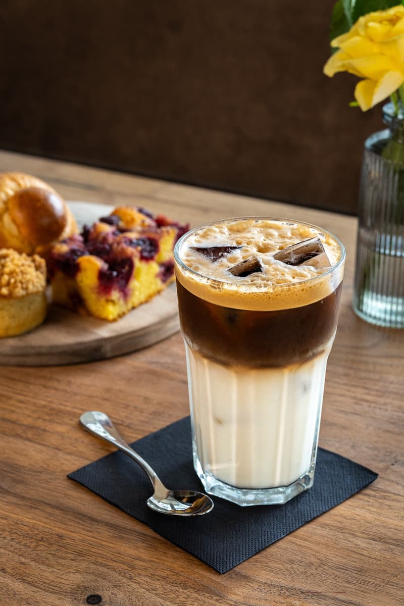 A glass of iced coffee or latte with a layer of foam on top, served on a wooden table along with a pastry or dessert item. There is also a vase with a yellow flower in the background.