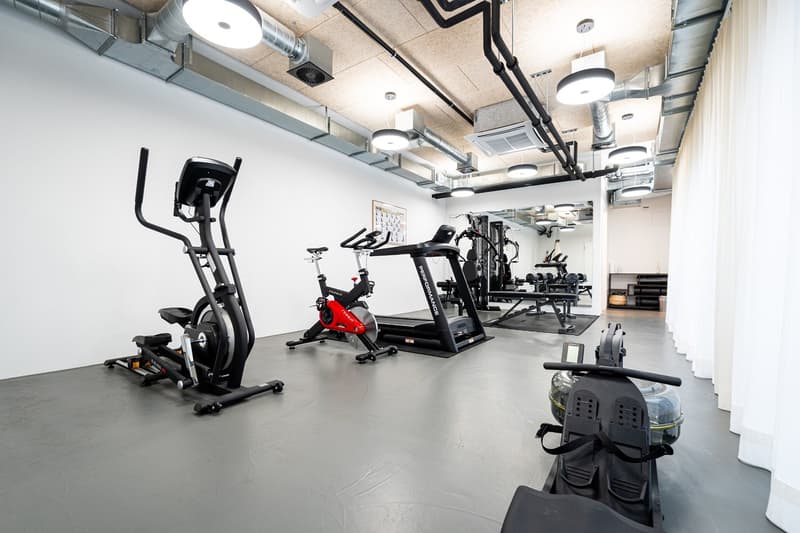 This image shows a modern, well-equipped gym space. The room features various fitness equipment such as elliptical machines, stationary bikes, and weight training machines. The ceiling has exposed ductwork and lighting fixtures, giving the space an indust