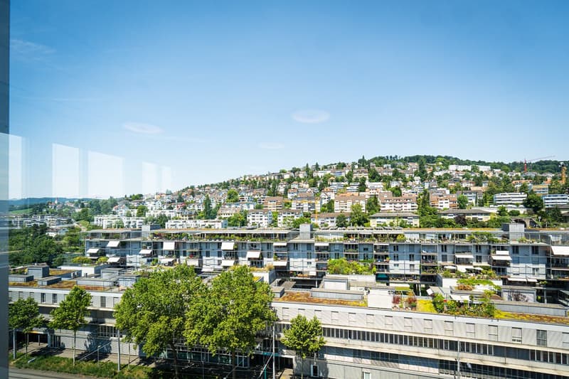 The image shows a panoramic view of a densely populated urban area with multi-story residential and commercial buildings nestled on a hillside. The buildings have balconies and terraces visible, and the cityscape is surrounded by lush greenery and a clear