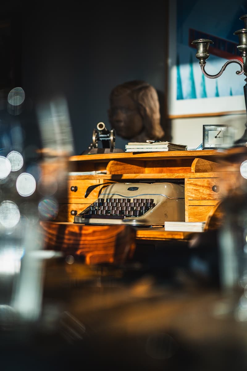 Vintage typewriter on a wooden desk, with a candle holder and other decorative items in the background. The room has a cozy, warm atmosphere with a fireplace visible.