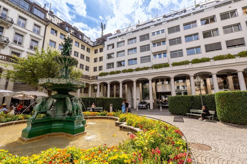 This image shows a large, multi-story building with a courtyard in the center. The building has a classical architectural style with arched entryways and decorative elements. There is a fountain with a green, ornate design in the center of the courtyard,