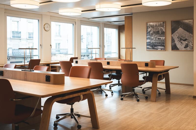 modern office room, long wooden desk, brown swivel chairs, large windows, wall clock, picture frames, wooden floor, lamps