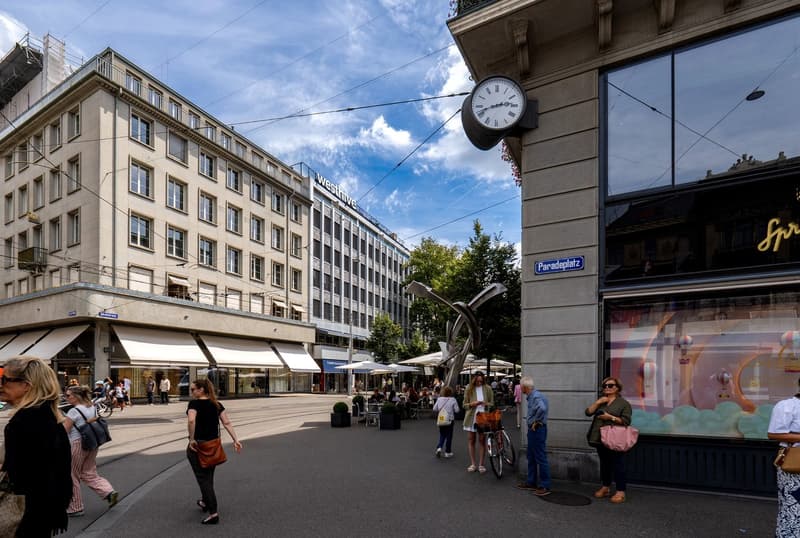 This image shows a busy city street with a mix of commercial and residential buildings. There is a large clock visible on one of the buildings, and people can be seen walking and sitting at outdoor cafes. The street has a lively, urban atmosphere with a m