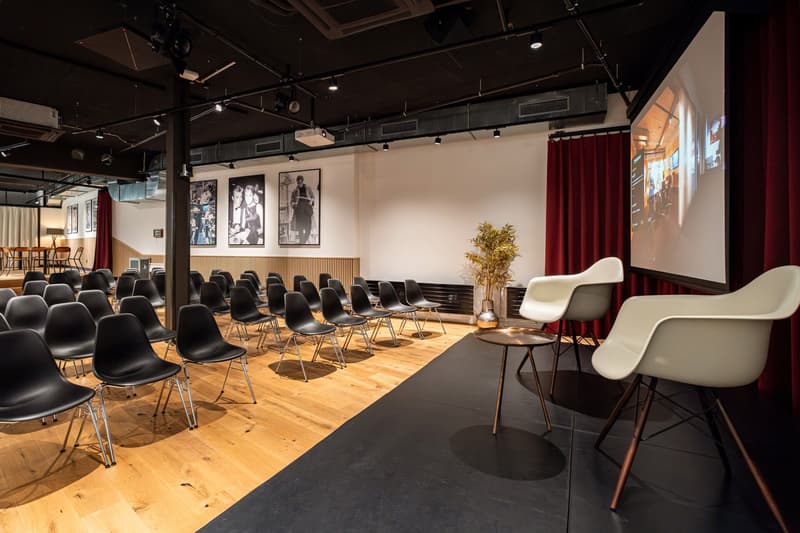 conference seating with black chairs, black floor in the front, white walls, projector screen, two armchairs, small table