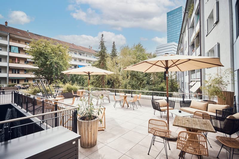 Outdoor patio area with tables, chairs, umbrellas, potted plants, and metal railings.