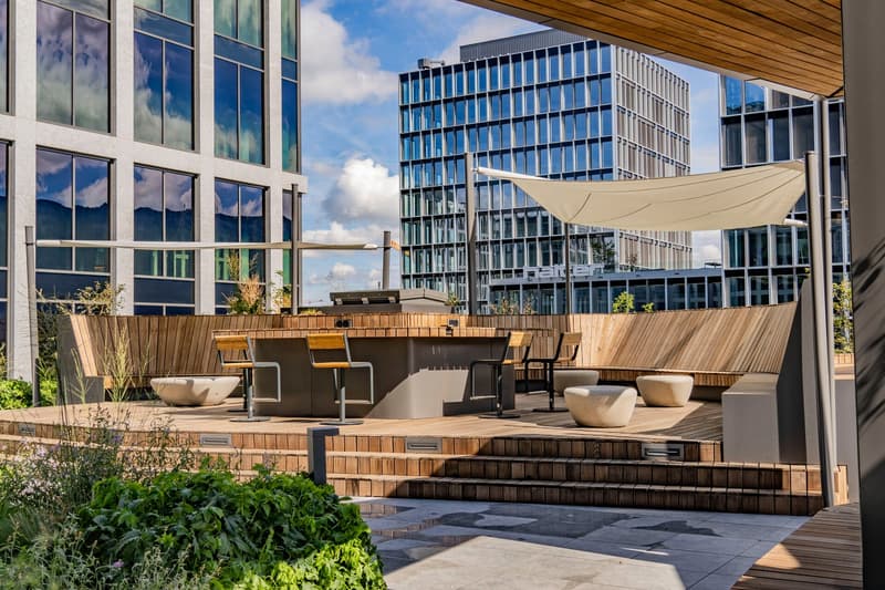 Wooden seating with a canopy and central bar counter, wooden benches, green plants, elevated wooden deck, modern buildings in the background