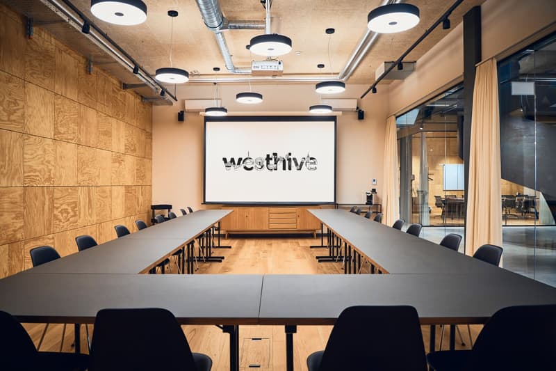 large rectangular conference room with wooden floors, long U-shaped tables, chairs, large screen projector, wooden wall paneling, and glass walls
