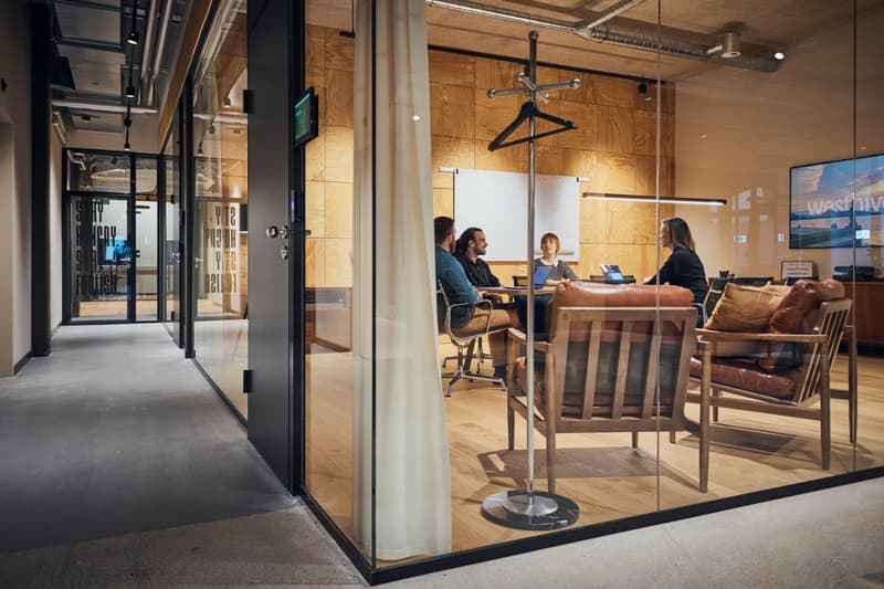 Modern office space with wooden walls, glass partitions, open floor plan, chairs and tables, glass doors, whiteboards, and monitors.