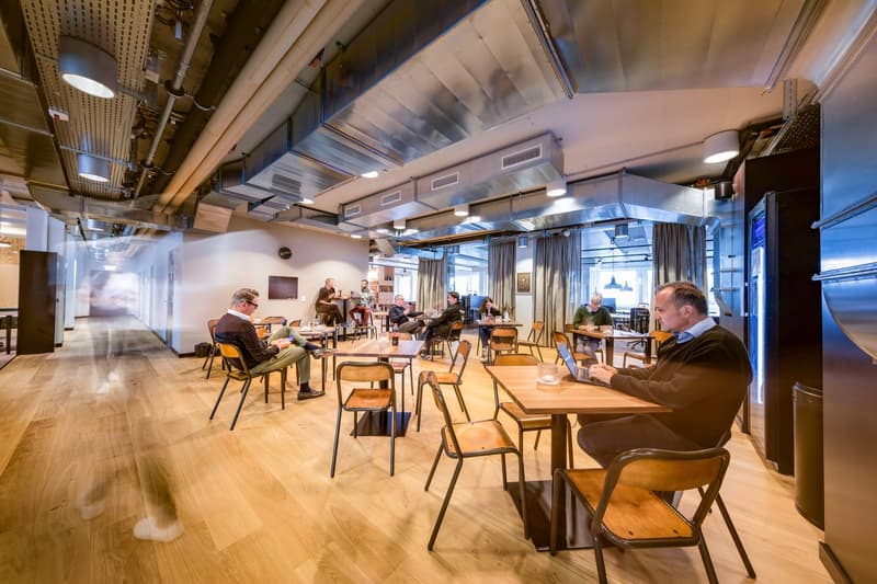 Wooden floor, metal chairs, rectangular tables, several people sitting, one person standing, ceiling lamps, metal structure visible