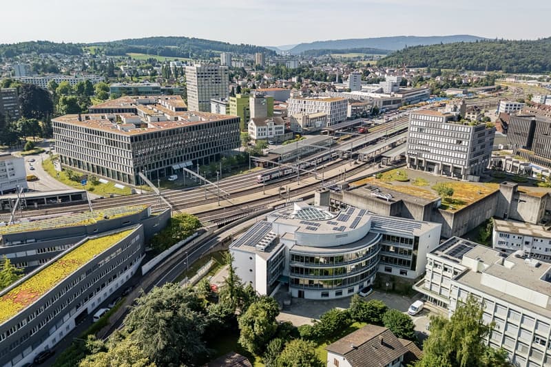 Aerial view of a city with a mix of modern high-rise buildings and older structures. The city appears to be situated in a hilly, forested area with a train station or transportation hub visible in the center of the image. There are several parking lots or