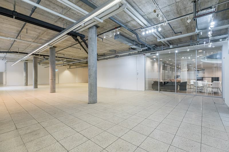 Large open office space with concrete ceiling, exposed pipes and ductwork, concrete floor tiles, and glass-enclosed meeting rooms or offices. The space appears to be a modern, industrial-style office environment.