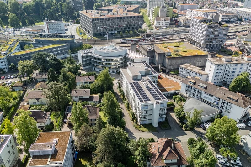 Urban aerial view of residential and commercial buildings, green rooftops, solar panels on some buildings, greenery, roads, and vehicles.