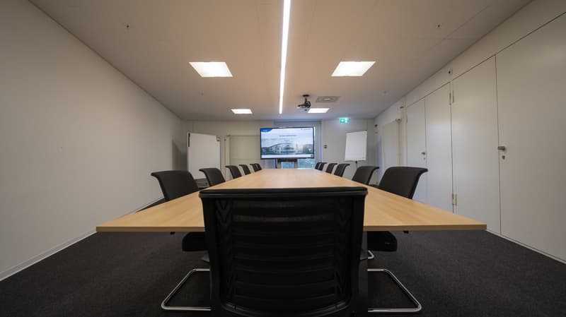long wooden table, black chairs, projector screen, ceiling lights, white walls