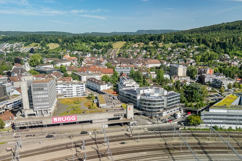 Aerial view of a city with a mix of modern and historic buildings, surrounded by lush green hills and forests. The city has a train station and parking facilities visible in the foreground.