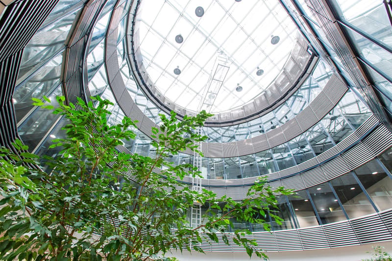 Large glass dome-shaped atrium with a curved, geometric steel frame structure. The atrium is filled with lush green plants and trees, creating a natural, indoor garden-like environment.