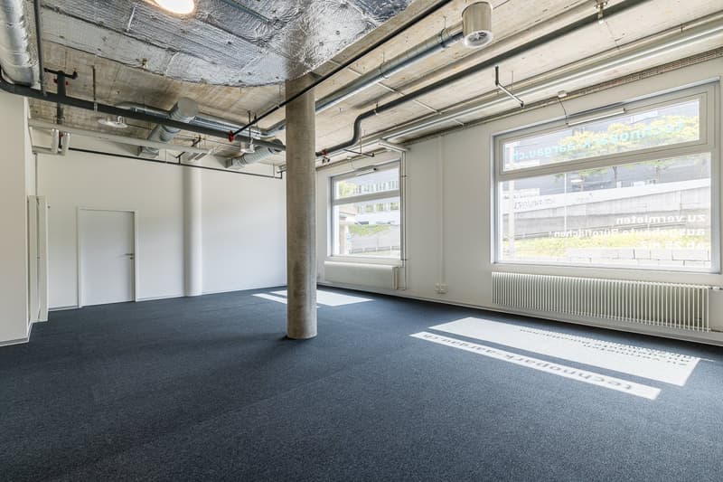Large open office space with exposed ceiling, concrete columns, and large windows overlooking the exterior. The space has a gray carpet floor and appears to be in a commercial or industrial building.