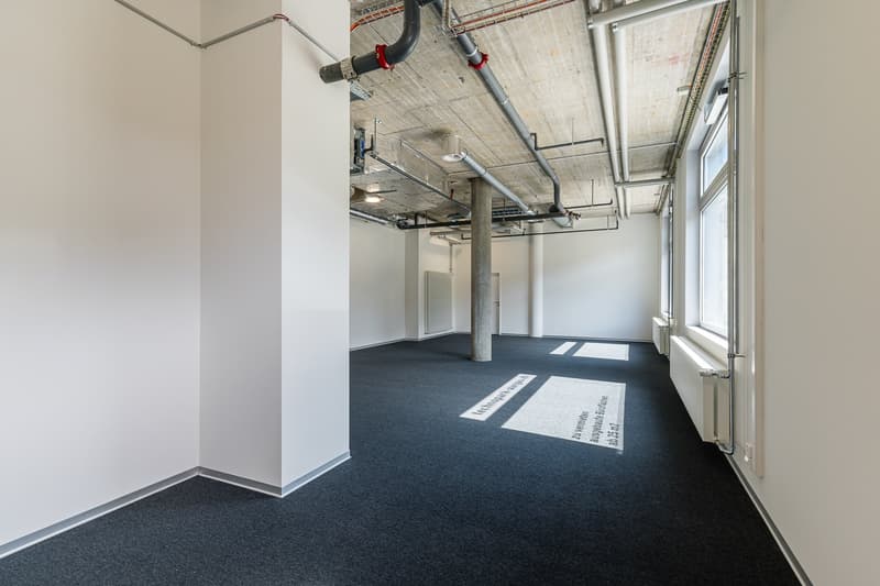 Large open office space with exposed ceiling, concrete walls, and dark carpeted flooring. The space has ample natural light from the windows and appears to be a commercial or industrial-style office environment.