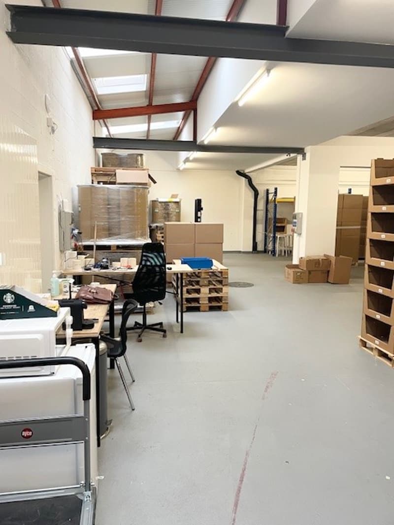 Warehouse with grey flooring, multiple pallets, office tables, storage boxes, ceiling lights, and an open space.