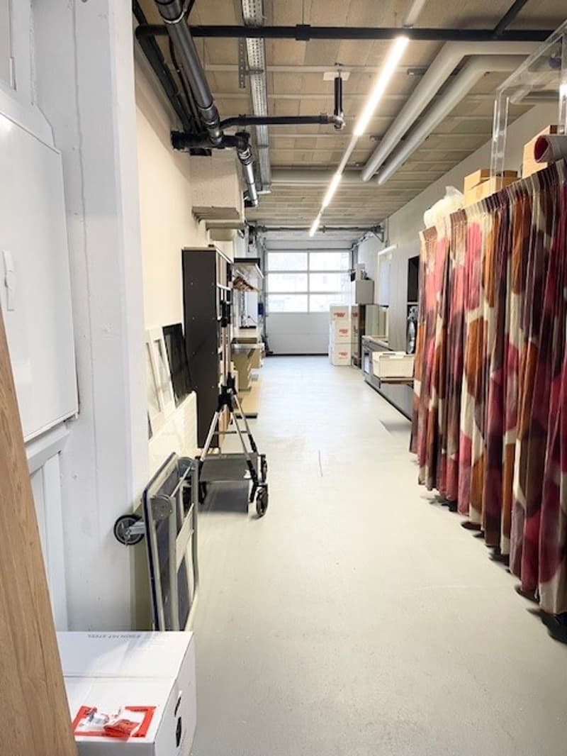 An indoor space with white walls, ceiling with pipes, rows of fabric hangings, boxes, a whiteboard, and a cabinet with items. The floor is white, and there are two windows with white frames. The room is empty with no furniture.