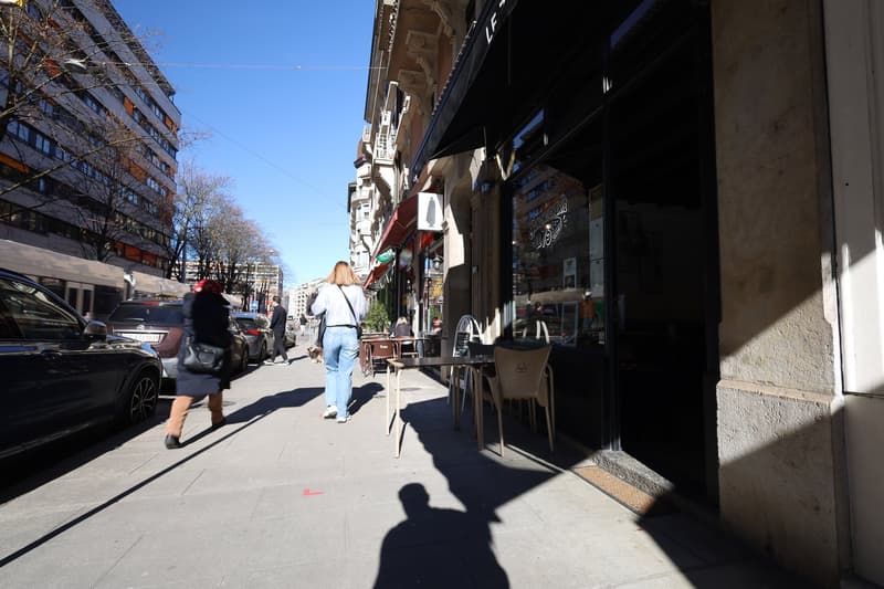 City street with a cafe on the right, people walking on the sidewalk, cars parked on the left, and buildings in the background.