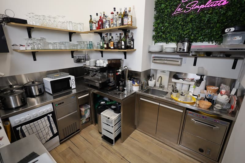 modern kitchen with stainless steel counters, gas stove, microwave, and sink. Bar with coffee machine, wine bottles, and shelves with glasses. Green wall decoration and neon sign.