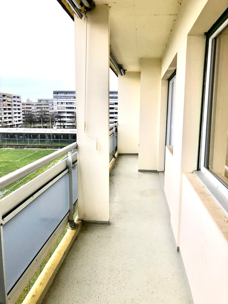 Empty balcony with metal railings, white walls, and a view of a grassy field and buildings
