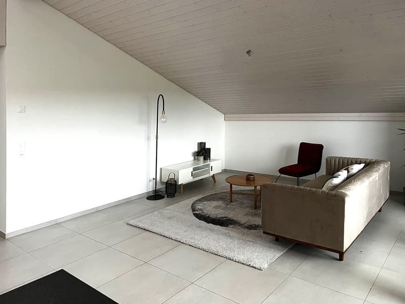 A white walled attic with a tile floor, a beige sofa, a red chair, a coffee table, and a floor lamp