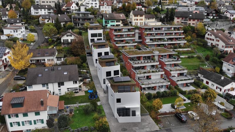 Modern houses in hilly area, with raised ground floor, several balconies, terraces, solar panels on roofs, parking places, surrounded by greenery