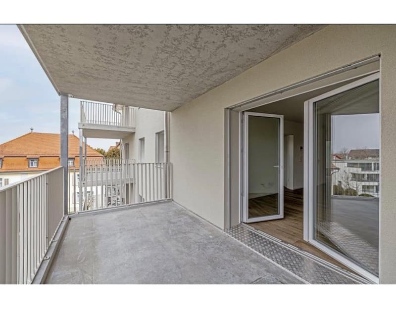 Open balcony with railings, sliding glass doors, and concrete floor, leading to a room with wooden floor and white walls.