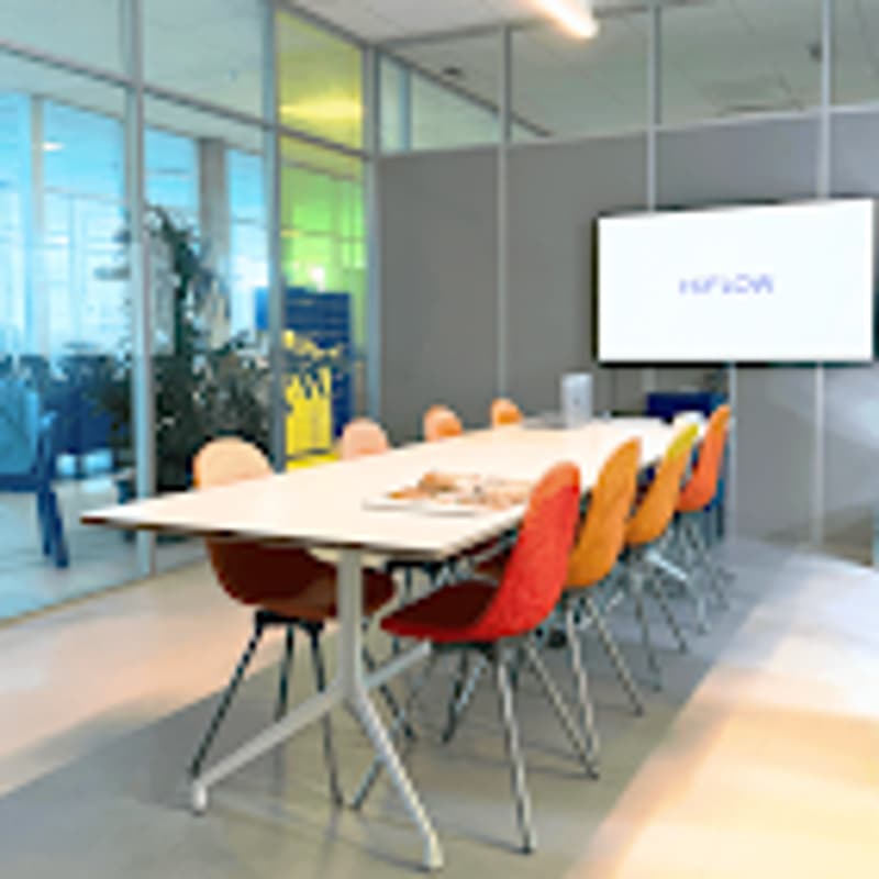 Empty meeting room with glass walls, long white table, orange and blue chairs, screen, whiteboard