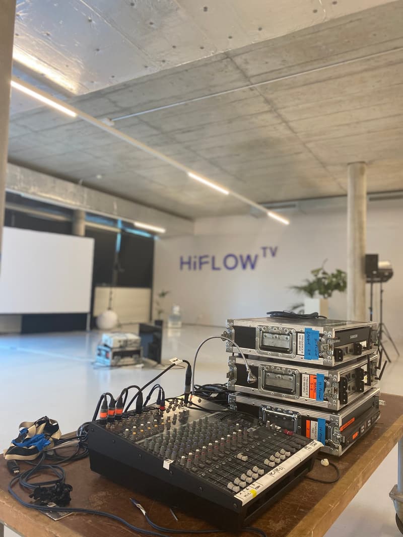 Empty room with white walls, grey ceiling, a tripod with camera, several electronic equipment on a table including a mixing console, and text that reads 'HiFLOW TV' on the wall.